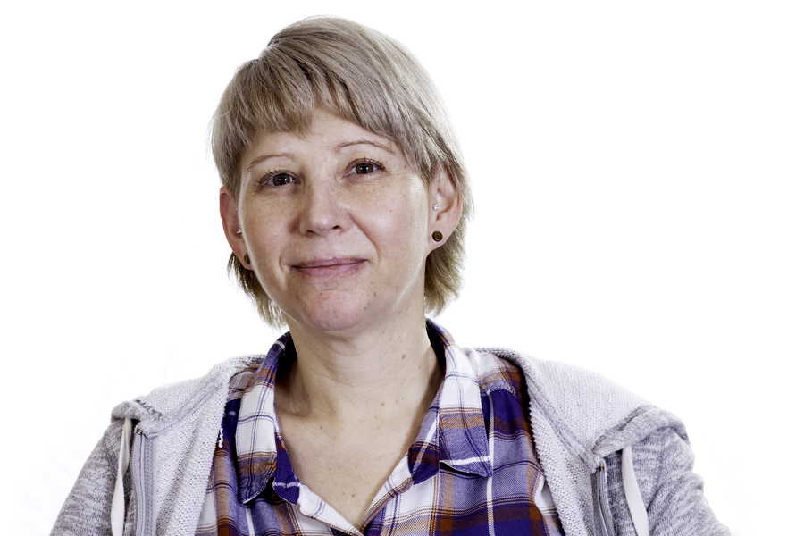 This Brian Charles Steel photo is a headshot of a white woman with short blonde hair.  She is wearing brown earrings and a grey sweater jacket over a blue, orange and white plaid shirt. She is centered in the frame, and the background is completely white.  She is lit in a Rembrandt style. 